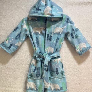 May include: A light blue fleece bathrobe with a hood, featuring a pattern of white polar bears, green trees, and blue foliage. The robe has a matching belt and pockets. The robe is designed for children.