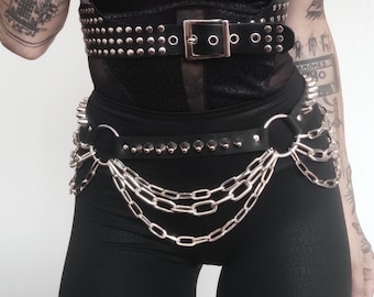 May include: A black leather harness with silver studs and a black leather belt with silver studs and chains. The harness has a leopard print bra underneath. The person is wearing black pants.