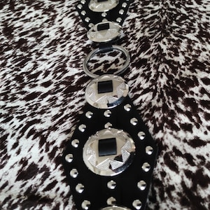 Peut inclure: Une ceinture en cuir noir avec des accents en concho argent&eacute;s. La ceinture a une rang&eacute;e de clous argent&eacute;s de chaque c&ocirc;t&eacute;. La ceinture est sur un fond &agrave; motifs blanc et brun.