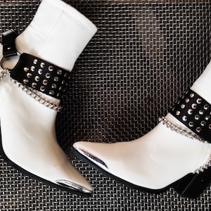 Puede incluir: Botas blancas de tobillo con punta puntiaguda y detalles metálicos plateados. Las botas tienen correas de cuero negro con tachuelas plateadas y cadenas plateadas.