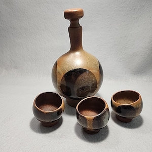 May include: A ceramic decanter set. The decanter is round, brown and tan, with a dark brown circular design. It has a stopper. Three small, matching cups are also included. The cups are round and have a similar brown and tan colour scheme.