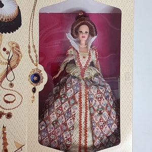 May include: A porcelain doll wearing a gold and red gown with a patterned design. The doll has brown hair and is wearing a pearl necklace. The doll is in a box with a gold and white design.