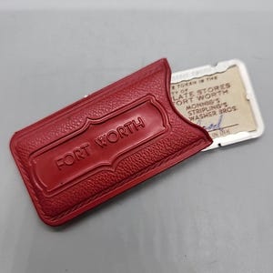 May include: A red leather case with the embossed text "FORT WORTH" holds a rectangular token. The token has text that reads "TOKEN IN THE CITY OF LATE STORES FORT WORTH MONNIG'S STRIPLING'S WASHER BROS."