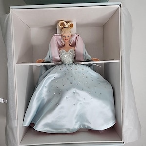 May include: A porcelain doll wearing a light blue gown with a pink shawl and a tiara. The gown is covered in sparkling rhinestones.