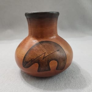 May include: A small, brown ceramic vase with a dark brown rim and a rounded body. The vase features a dark brown bear design with an arrow. The bear design is on the front of the vase.