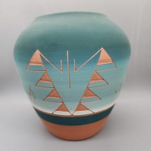 May include: A turquoise ceramic vase with a geometric design. The vase features a gradient of colors, transitioning from a darker teal at the top to a lighter blue in the middle. The base is a terracotta color, with a dark green band separating it from the blue. The design is a series of triangles.
