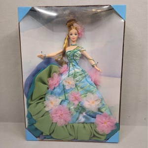 Puede incluir: Una muñeca rubia en una caja que lleva un vestido largo hasta el suelo con un estampado de acuarela azul y verde, adornado con flores de tela rosa, detalles de cuentas verdes y un dobladillo verde con volantes. Peinado recogido con flor rosa.