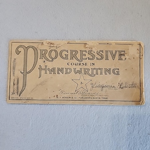 以下が含まれることがあります： 「Progressive Course in Handwriting」と題された、古い手書き指導書。表紙はベージュ色で、黒い文字と星のグラフィックが特徴です。「Virginia Roberts」の名前と、テキサス州オースティンにある出版社の住所が記載されています。