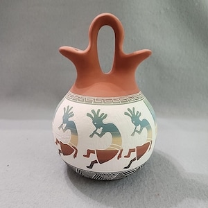 Könnte beinhalten: Ein handgefertigtes, terrakottafarbenes Keramikgefäß mit einem markanten Griff- und Ausgussdesign. Der weiße Körper zeigt Kokopelli-Figuren in Blau-, Gelb- und Brauntönen mit geometrischen Mustern. Ein einzigartiges Kunstwerk aus dem Südwesten.