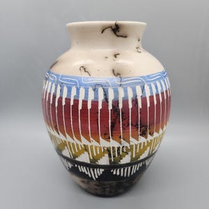 May include: A ceramic vase with a cream-colored base featuring black horsehair-style markings. The body displays horizontal bands of blue, red, orange, and yellow, overlaid with a repeating white geometric pattern and vertical white lines.