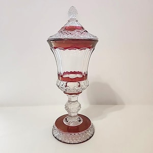 May include: A clear glass decorative jar with a ruby red rim and accents. The jar has a lid and a pedestal base. The base and lid have a diamond pattern. The jar is approximately 30 cm tall.