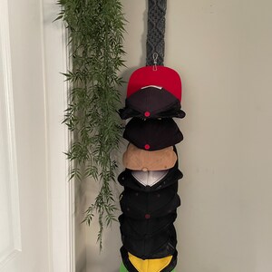 May include: A macramé hat holder with a wooden ring at the top, holding a stack of baseball caps. The caps are various colours, including red, black, tan, and green. The holder is hanging on a wall next to a hanging green plant.