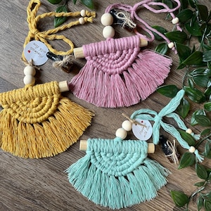 May include: Three macrame car air fresheners with wooden dowels and tassels. The air fresheners are in yellow, pink, and light blue. Each air freshener has a small bottle of essential oil attached to it with a string. The tag on the yellow air freshener reads "Handmade with love".