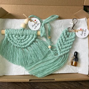 May include: A set of two macrame wall hangings in a light green colour, with a wooden dowel and fringe. The set includes a small macrame keyring with a silver key ring. The hangings are packaged in a brown cardboard box with white tissue paper. The hangings have tags that say "Handmade with love".
