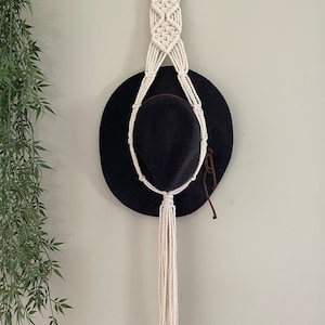 May include: A cream macrame hat holder with a black hat. The holder is suspended from a wooden ring and features a woven design with a long tassel. The hat is a wide-brimmed style, suitable for a variety of head sizes.
