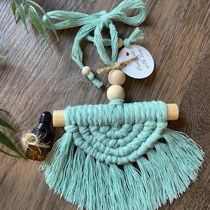 May include: A light green macrame wall hanging with wooden dowels and a small bottle of essential oil. The hanging has a fringe and a tag that says "Handmade with love".