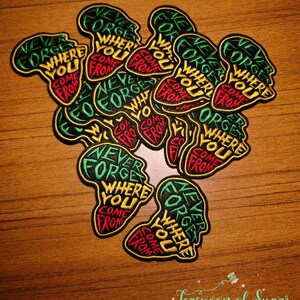 May include: Embroidered patches in the shape of Africa, featuring the text "Never Forget Where You Come From" in green, yellow, and red. The patches are black with yellow borders, scattered on a wooden surface.