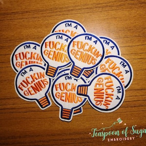 May include: A pile of white, orange, and blue embroidered iron-on patches shaped like light bulbs. Each patch says "I'm a fuckin' genius".