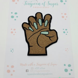 May include: Embroidered patch featuring a brown hand with black outlines and light blue fingernails. The patch is on a white card with the text "Teaspoon of Sugar EMBROIDERY" and social media information.