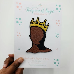 Pode incluir: Um remendo bordado marrom de uma pessoa usando uma coroa dourada. O remendo está em um cartão branco com pontos rosa e azuis. O cartão tem o texto "Teaspoon of Sugar Embroidery" e "Made with a Teaspoon of Sugar".