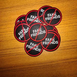 May include: A collection of black circular patches with the words "FAKE FRIENDS" in white, crossed out with a red line. The patches have a red border. The patches are on a wooden surface.