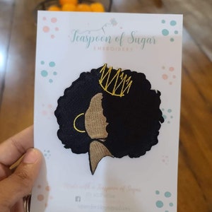 May include: Black and gold embroidered patch of a woman with an afro and a crown. The patch is on a white card with pink and blue polka dots. The card says "Teaspoon of Sugar Embroidery".