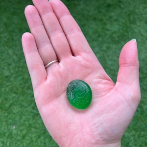 May include: A small, round, emerald green glass stone with an etched spiral design. The stone is held in an open hand against a backdrop of green grass.