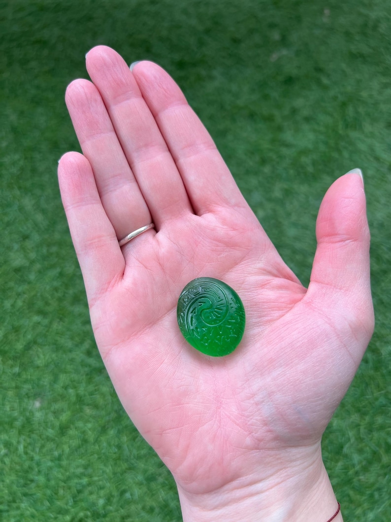 May include: A small, round, emerald green glass stone with an etched spiral design. The stone is held in an open hand against a backdrop of green grass.