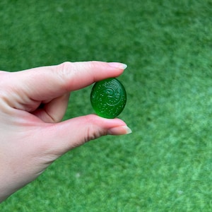 May include: A small, oval-shaped, translucent green glass bead with an engraved floral design. The bead is held against a backdrop of green grass. The bead is approximately 1 inch in length.