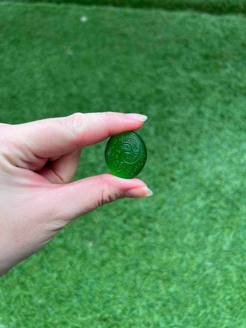 May include: A small, oval-shaped, translucent green glass bead with an engraved floral design. The bead is held against a backdrop of green grass. The bead is approximately 1 inch in length.