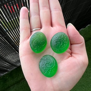 May include: Three oval, translucent green stones with carved spiral designs. The stones are held in an open hand, showcasing their vibrant color and intricate patterns. The background is blurred, focusing attention on the gemstones.