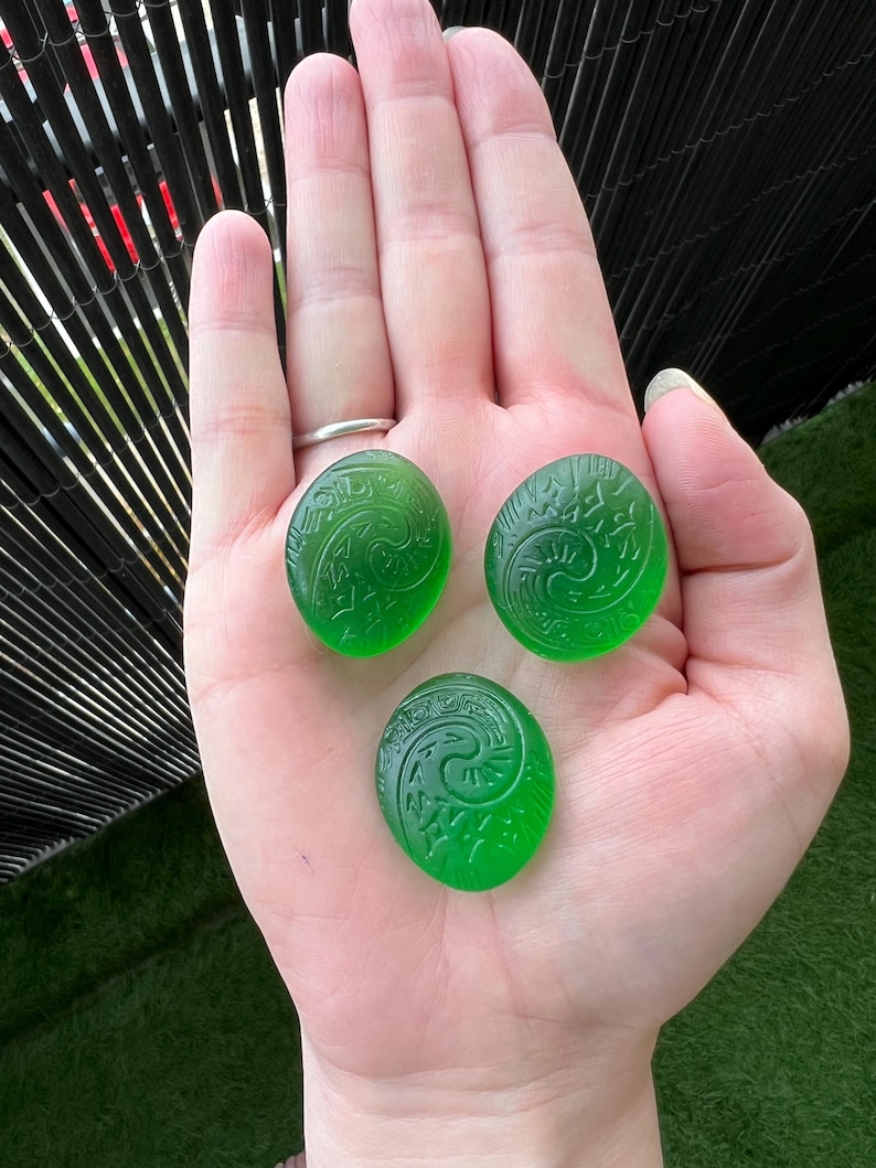 May include: Three oval, translucent green stones with carved spiral designs. The stones are held in an open hand, showcasing their vibrant color and intricate patterns. The background is blurred, focusing attention on the gemstones.
