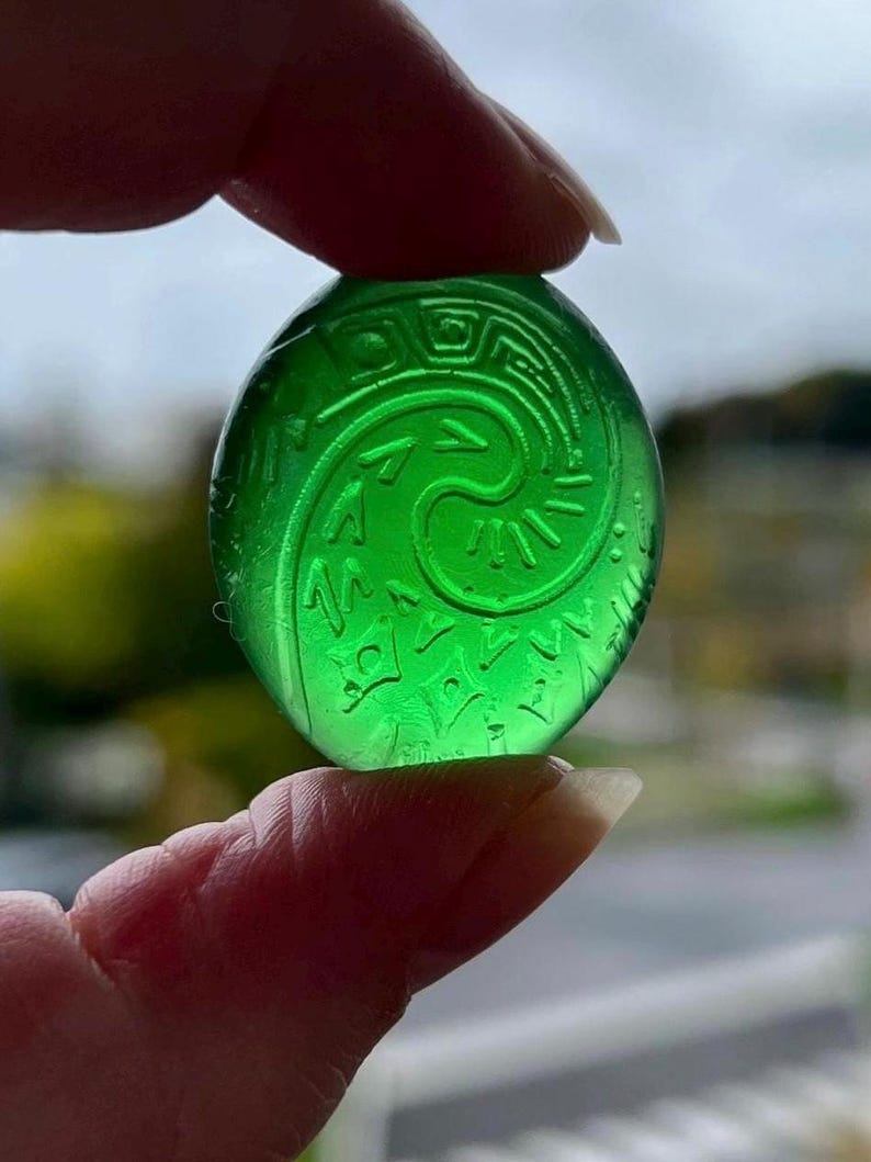 May include: A translucent, round, green glass pendant with an engraved spiral and geometric design. The pendant is held between fingers, showcasing its intricate details and vibrant color. The background is blurred, emphasizing the pendant.
