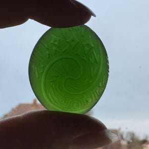 Pode incluir: Um pingente oval verde translúcido com um design espiral e geométrico gravado. O pingente é segurado entre os dedos, mostrando seus detalhes intrincados e sua cor vibrante. O design lembra a arte antiga.