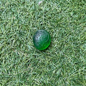 May include: A round, translucent green glass bead with an engraved spiral design. The bead is resting on a bed of green artificial grass. The bead's design is a swirling pattern.
