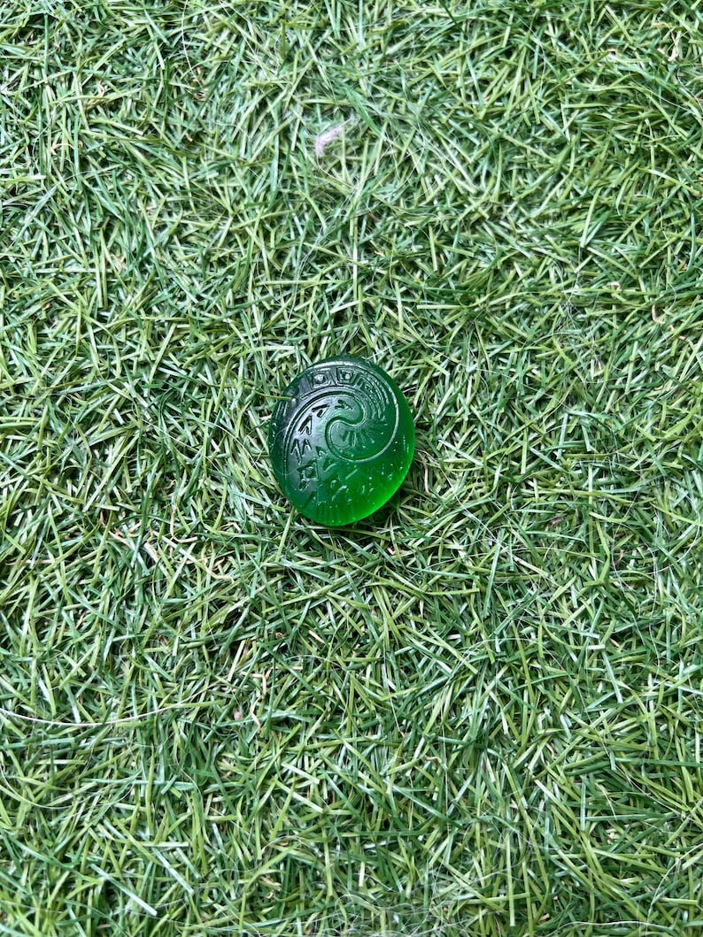 May include: A round, translucent green glass bead with an engraved spiral design. The bead is resting on a bed of green artificial grass. The bead's design is a swirling pattern.