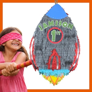 May include: A grey rocket-shaped piñata with red and silver details, a blue top, and colourful flames. The piñata is decorated with the name "JAMISON" and "1st". A child wearing a blindfold is holding a bat, preparing to hit the piñata.