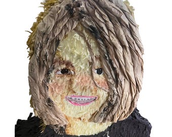 May include: A piñata shaped like a human head and shoulders, featuring light brown paper fringe hair, a cream-colored face with painted eyes, a pink mouth with visible teeth, and a black paper fringe base representing clothing or a shirt.