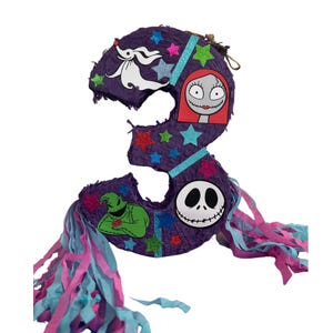 Puede incluir: Una pi&ntilde;ata con forma de n&uacute;mero tres de color morado decorada con estrellas brillantes y recortes de personajes que incluyen un perro fantasma blanco, una figura pelirroja, un monstruo verde y una calavera blanca con tiras de papel.