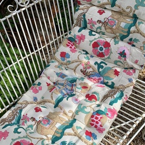 May include: A white metal garden bench with a floral patterned cushion. The cushion has a cream background with pink, blue, and green floral designs. The cushion is quilted and has a tufted design.