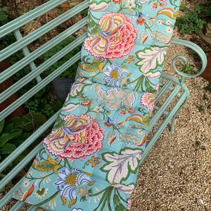 Puede incluir: Un coj&iacute;n de banco rectangular con un estampado floral sobre un fondo azul claro. Presenta grandes flores rosas, amarillas y verdes. El coj&iacute;n est&aacute; dise&ntilde;ado para un banco de metal. El dise&ntilde;o incluye hojas grandes y patrones florales detallados.