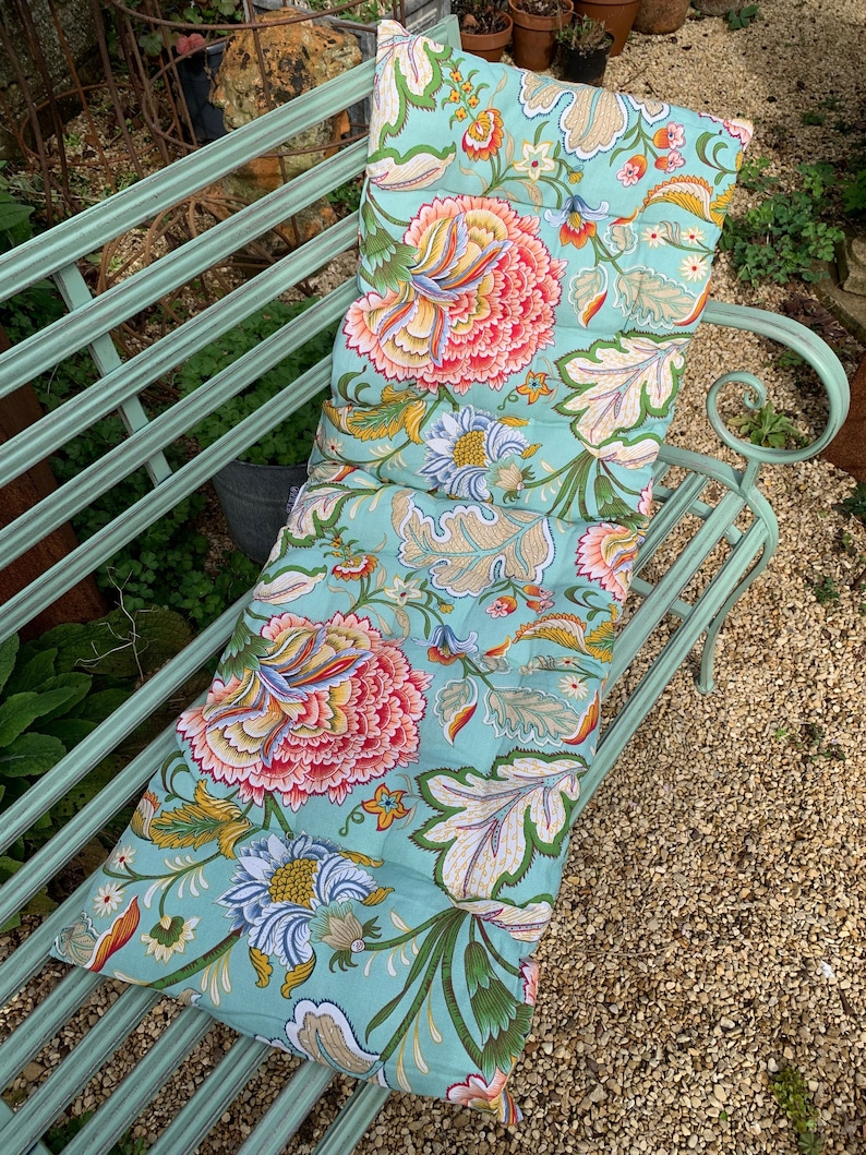 Puede incluir: Un coj&iacute;n de banco rectangular con un estampado floral sobre un fondo azul claro. Presenta grandes flores rosas, amarillas y verdes. El coj&iacute;n est&aacute; dise&ntilde;ado para un banco de metal. El dise&ntilde;o incluye hojas grandes y patrones florales detallados.