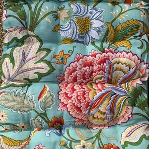Puede incluir: Un coj&iacute;n con estampado floral con grandes flores coloridas en tonos rojos, amarillos, azules y blancos sobre un fondo verde azulado claro. El dise&ntilde;o incluye hojas y tallos detallados, creando un aspecto vibrante y decorativo. El coj&iacute;n tiene una apariencia acolchada.