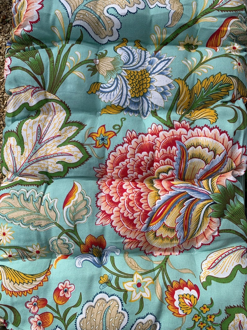 Puede incluir: Un coj&iacute;n con estampado floral con grandes flores coloridas en tonos rojos, amarillos, azules y blancos sobre un fondo verde azulado claro. El dise&ntilde;o incluye hojas y tallos detallados, creando un aspecto vibrante y decorativo. El coj&iacute;n tiene una apariencia acolchada.