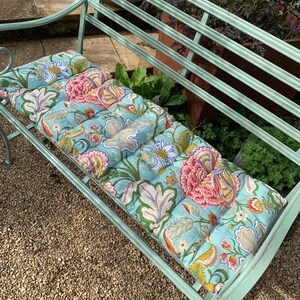 Puede incluir: Un banco de jard&iacute;n de metal verde menta con un coj&iacute;n estampado floral. El coj&iacute;n presenta un fondo azul claro con grandes dise&ntilde;os de flores rosas, naranjas y amarillas, junto con follaje verde y beige. El banco est&aacute; al aire libre sobre una superficie de grava.