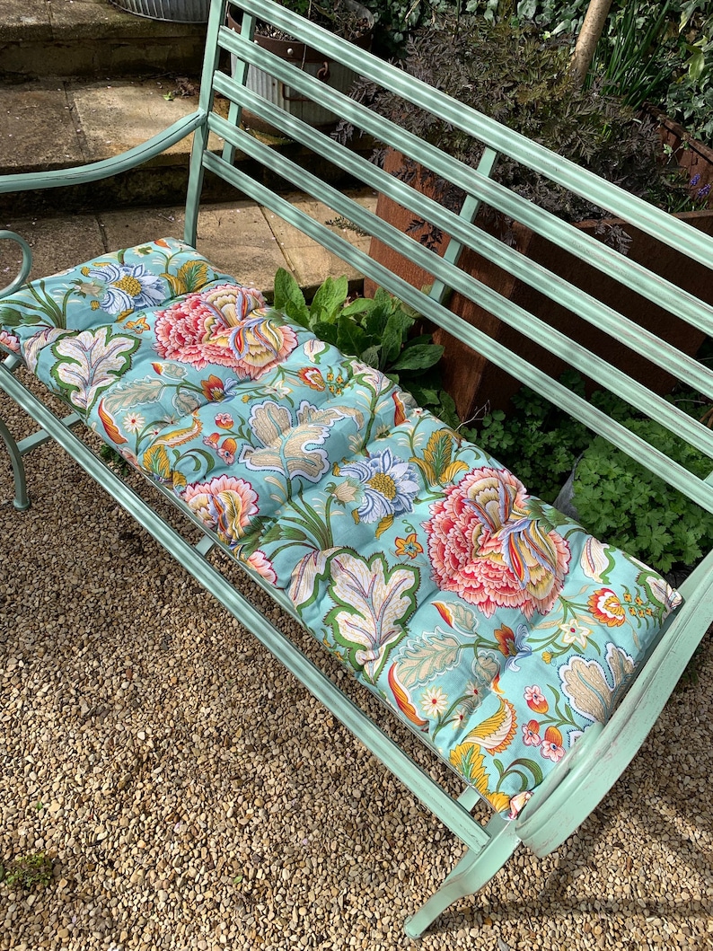 Puede incluir: Un banco de jard&iacute;n de metal verde menta con un coj&iacute;n estampado floral. El coj&iacute;n presenta un fondo azul claro con grandes dise&ntilde;os de flores rosas, naranjas y amarillas, junto con follaje verde y beige. El banco est&aacute; al aire libre sobre una superficie de grava.