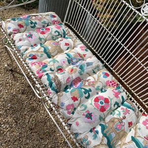 May include: White metal garden bench with a floral patterned cushion. The cushion has pink, red, green, and gold accents. The cushion is made of fabric with a quilted design.