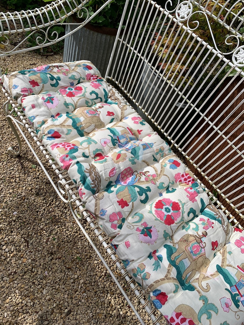 May include: White metal garden bench with a floral patterned cushion. The cushion has pink, red, green, and gold accents. The cushion is made of fabric with a quilted design.