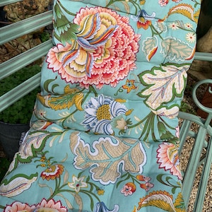 Puede incluir: Un coj&iacute;n de banco con estampado floral en tonos turquesa, coral, amarillo y verde. El coj&iacute;n presenta un dise&ntilde;o floral detallado y acolchado. Se encuentra sobre un banco de metal verde claro, sugiriendo su uso en exteriores.