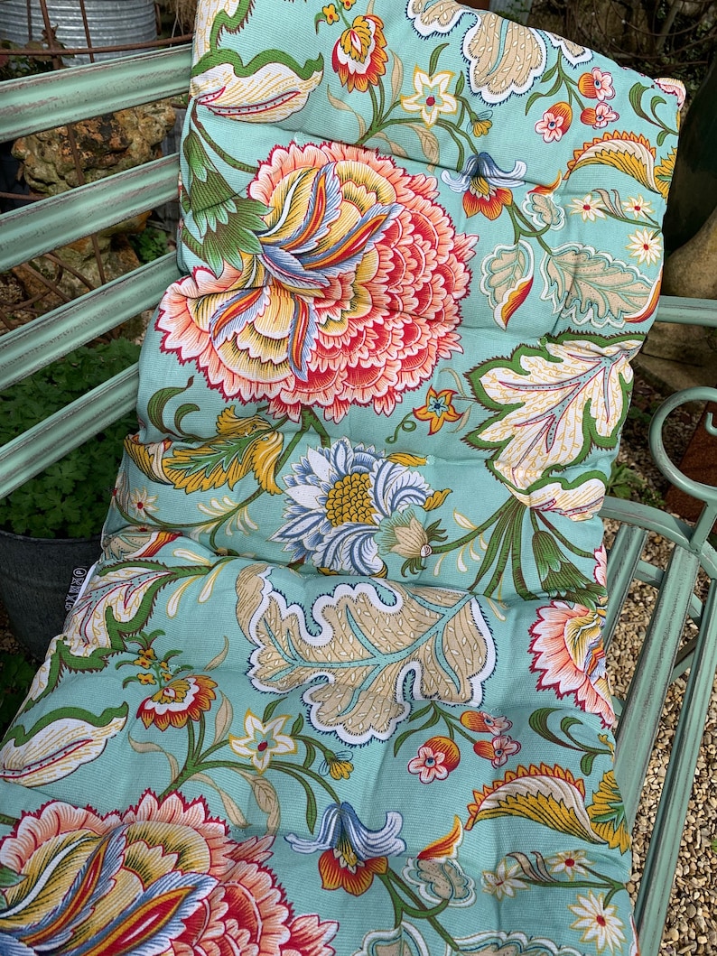 Puede incluir: Un coj&iacute;n de banco con estampado floral en tonos turquesa, coral, amarillo y verde. El coj&iacute;n presenta un dise&ntilde;o floral detallado y acolchado. Se encuentra sobre un banco de metal verde claro, sugiriendo su uso en exteriores.